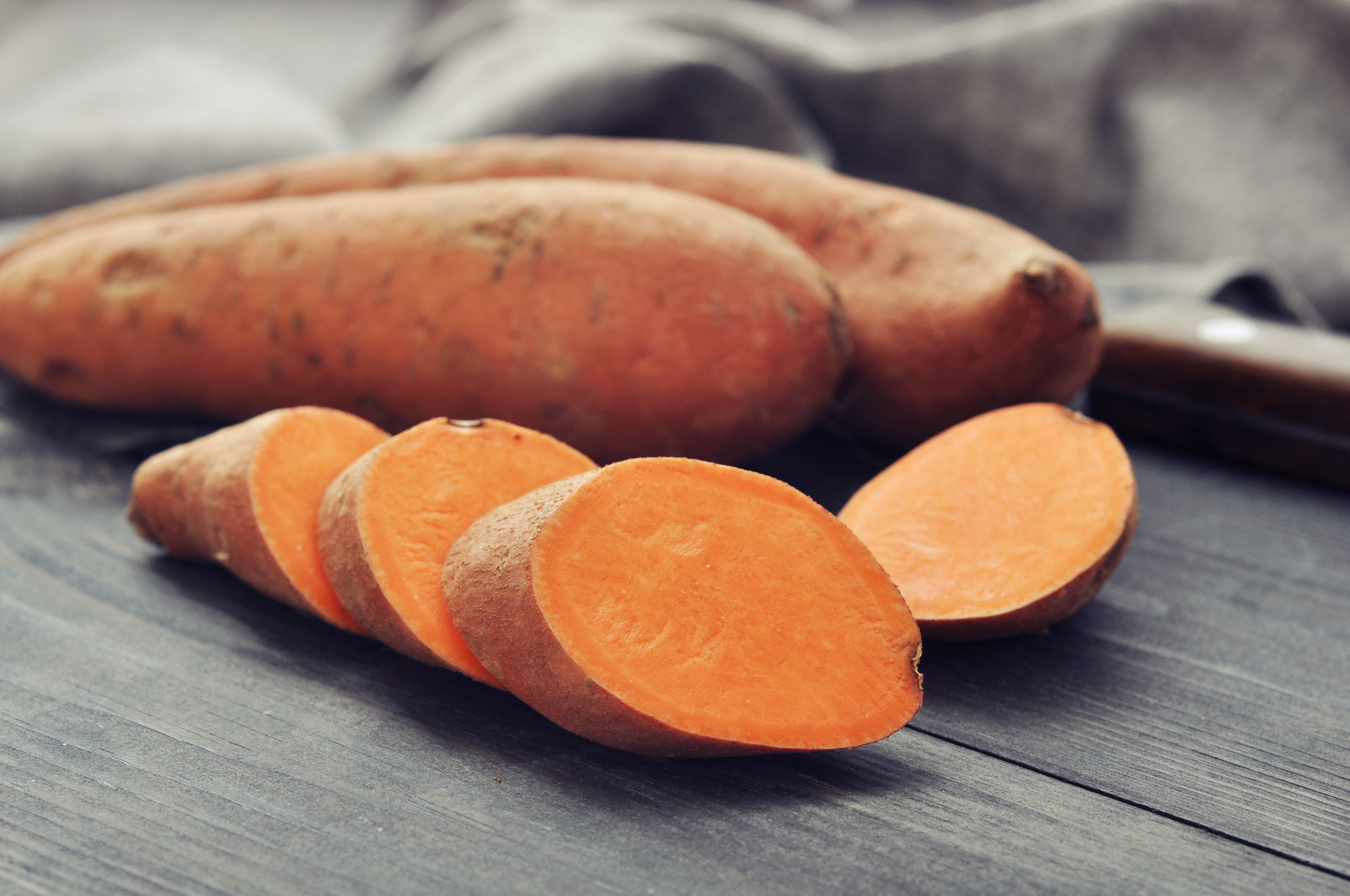 Raw sweet potatoes resting beside a kitchen knife on a dark wooden chopping board, ready for preparation